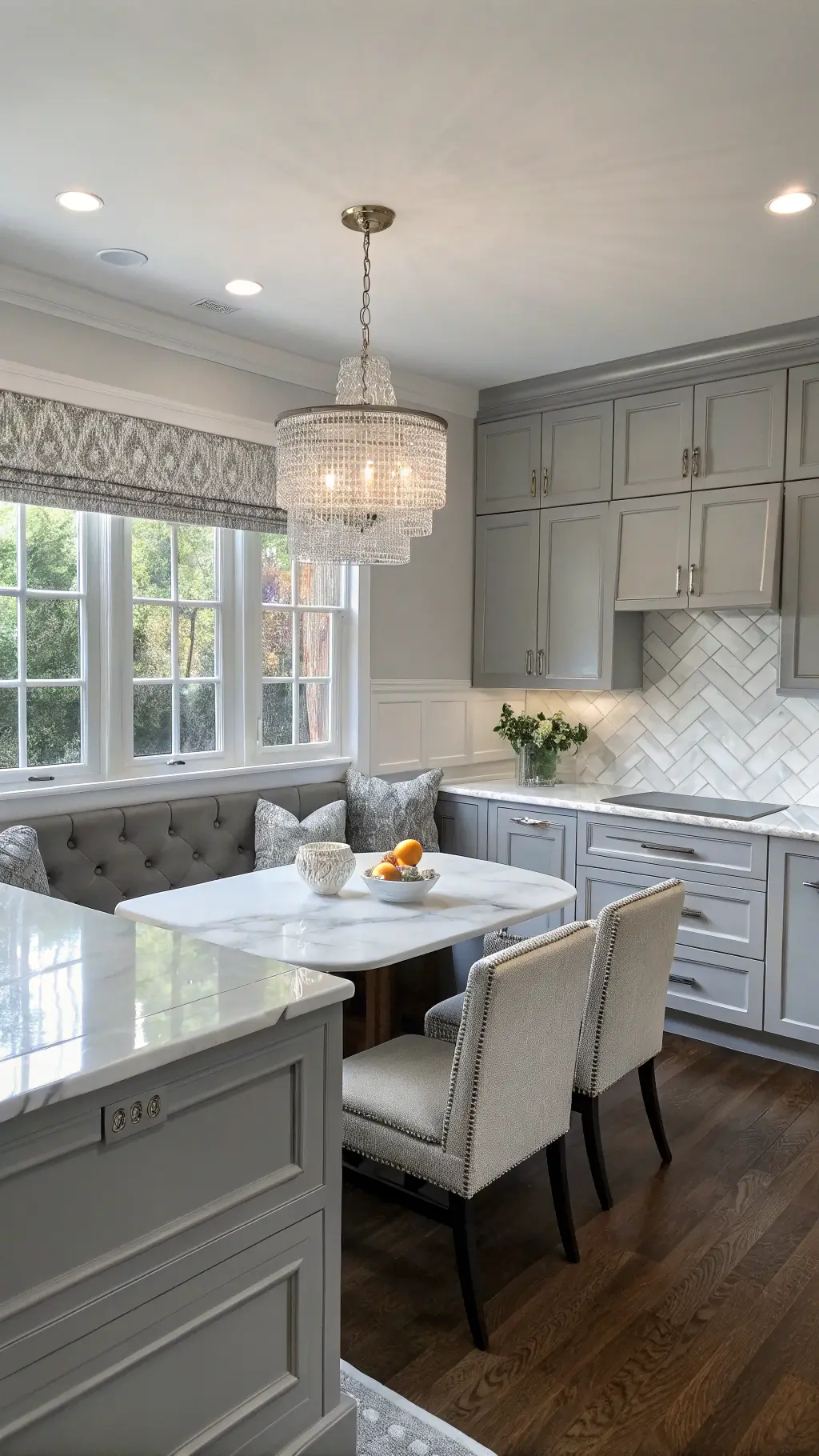 Elegant 16x20ft L-shaped, transitional grey kitchen with white marble countertops, grey herringbone backsplash, medium raised-panel cabinets and polished nickel pulls, breakfast nook, illuminated by soft mid-morning light through roman shades and crystal chandelier over island, showcasing full room depth.