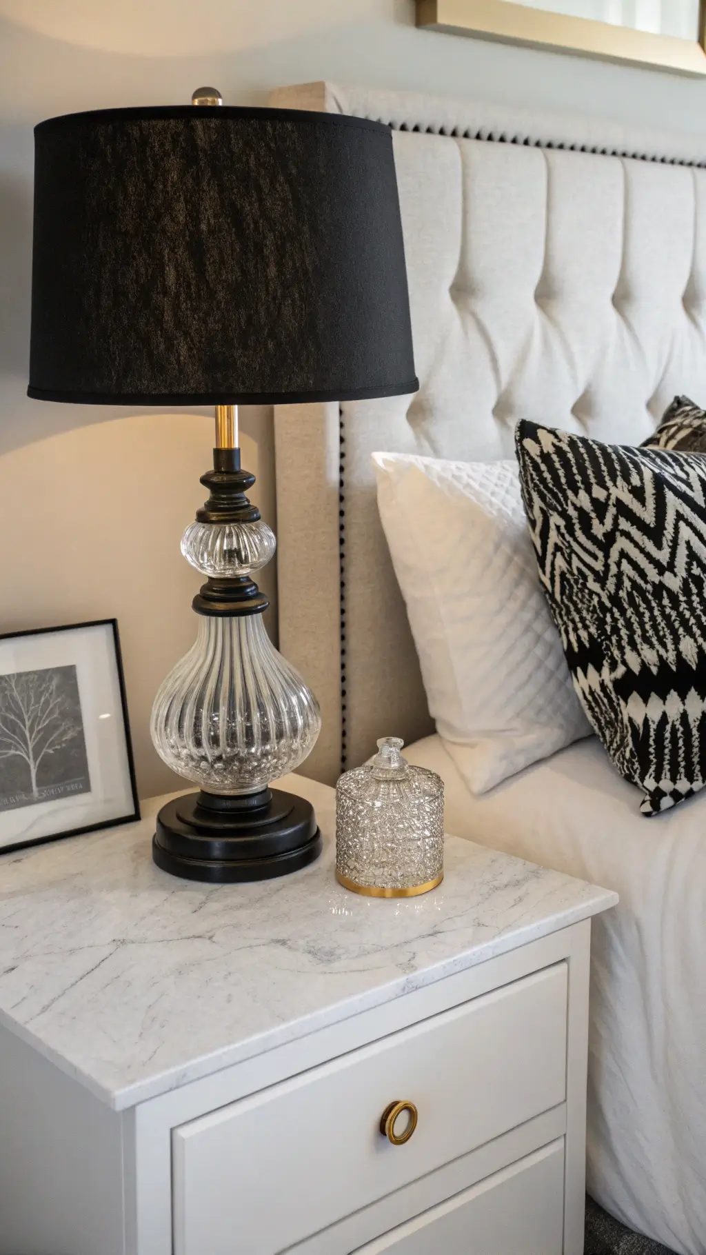 Late afternoon light highlighting styled white marble-top nightstand with black ceramic lamp, crystal carafe, and picture frame against white linen headboard with patterned pillows.