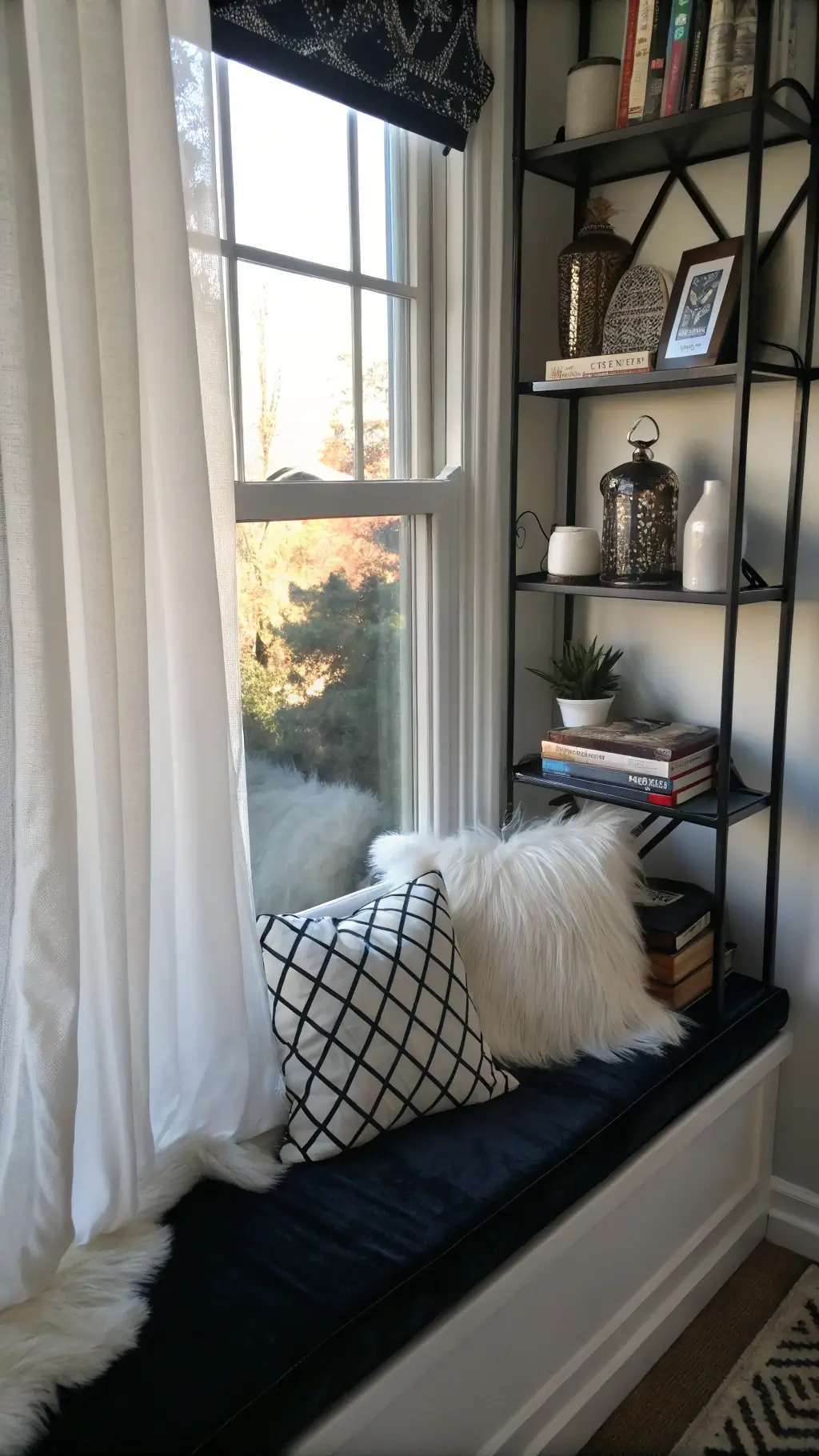 Dramatic chiaroscuro-lit window-side reading nook with a black velvet seat cushion, white faux fur and geometric pillows on floating black shelves displaying white pottery and art books framed by gauzy white curtains filtering afternoon light.