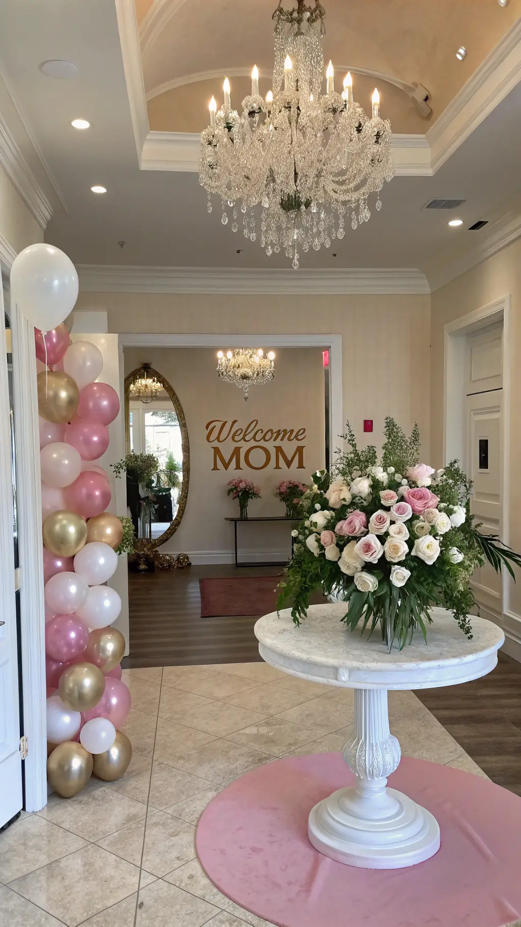 Stunning 10x12ft foyer with high ceilings, lit by natural light and a crystal chandelier, featuring white marble round table with a floral arrangement, mirror 'Welcome Mom' sign, blush pink carpet runner and metallic balloon bouquets on each side.